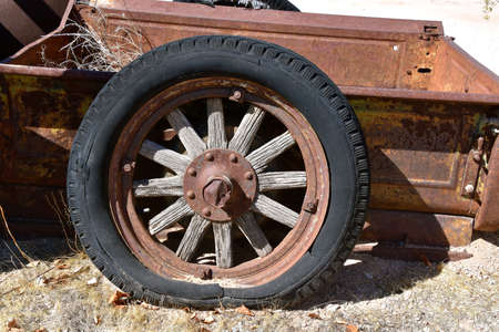 A very old wheel with wooden spokes, metal rim, and a rotten flat tire belong to a rusty box of a pickup or truckの写真素材