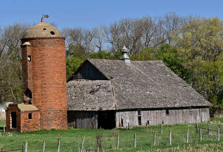 An unique hip roofed dairy barn and brick silo stand abandoned and in a deteriorating state.の写真素材