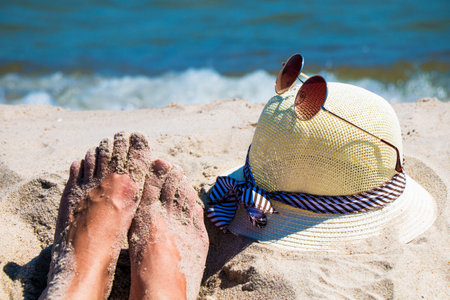 Close up of woman's feet and hat with sunglasses on sandy beach with blue sea - relaxation, holidaysの写真素材