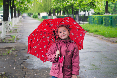 cheerful little girl with red umbrella in jacket stands in the middle of the sidewalk in the parkの写真素材