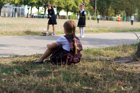 Schoolgirl girl in a sundress in a cage sitting on the grass looking at children with  skateboard outdoorsの写真素材