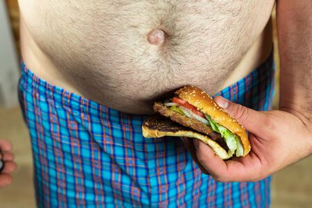 Man holding delicious and juicy burger against his fat belly. Anti fast food and time for a diet concept.の写真素材