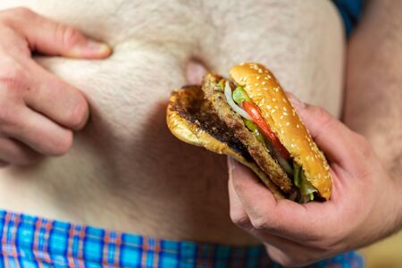 Man holding delicious and juicy burger against his fat belly. Anti fast food and time for a diet concept.の写真素材