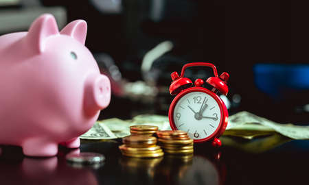 A red alarm clock on a dark colored table with small piles of coins, a pink piggy bank and US dollar bills in the background.の写真素材