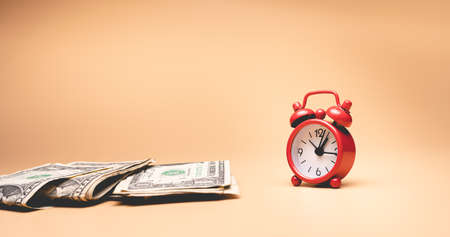 Time is money concept. Red alarm clock and dollar banknotes on beige background.の写真素材