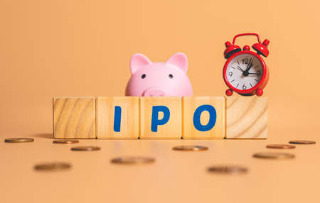 The word IPO is written on wooden cubes next to a piggy bank and a red alarm clock on a beige background.の写真素材