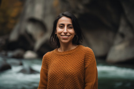 Portrait of smiling woman in sweater looking at camera while standing near riverの素材