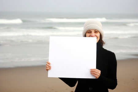 Young woman holding a blank sheet of paper on the beach in winterの素材