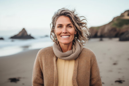 Portrait of happy mature woman smiling at camera on beach during autumn dayの素材