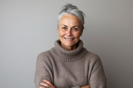 Portrait of smiling senior woman with crossed arms looking at camera. Mature woman with gray hair standing against gray background.の素材