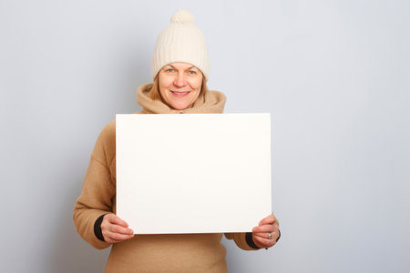 Smiling middle aged woman in winter clothes holding a white sheet of paperの素材