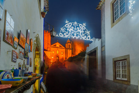 OBIDOS, PORTUGAL - DECEMBER 27, 2017: View of an alley in the old town, with local businesses, Saint James church, and Christmas decorations, in Obidos, Portugalのeditorial素材