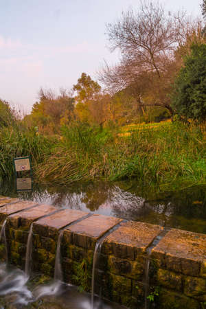 Sunset view of trees, fall foliage, the Kesalon Stream, and a trilingual warning sign, in En Hemed...