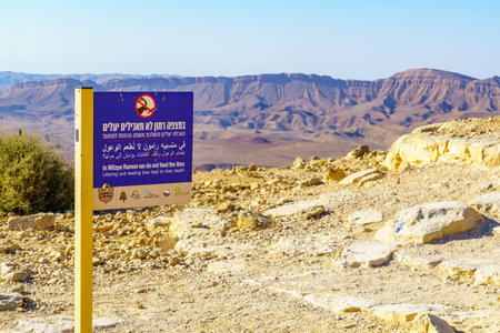 Mitzpe Ramon, Israel - August 11, 2021: Trilingual sign calling not to feed the ibex, and landscape of Makhtesh (crater) Ramon, in the Negev Desert, Southern Israelのeditorial素材