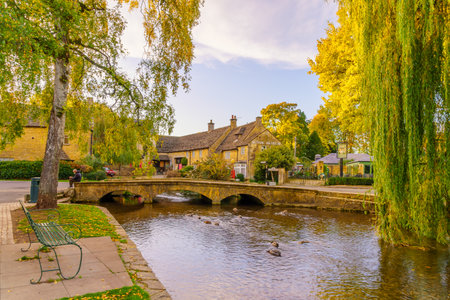 Bourton-on-the-Water, UK - October 17, 2022: Sunset scene of typical houses, the river Windrush, locals and visitors, in the village Bourton-on-the-Water, the Cotswolds region, England, UKのeditorial素材