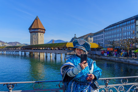 Lucerne, Switzerland - February 20, 2023: Participant in costume takes a selfie, crowd and city monuments in the background, part of the Fasnacht Carnival, in Lucerne (Luzern), Switzerlandのeditorial素材