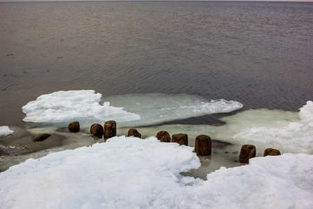 Jetty Pillars and Rocks Covered witn Snow in a Cloudy Winter Day with a Few Sun Beams Shining on Them and a Sea in a Backgroundの写真素材