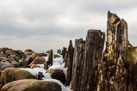 Jetty Pillars and Rocks Covered witn Snow in a Cloudy Winter Day with a Few Sun Beams Shining on Them and a Sea in a Backgroundの写真素材