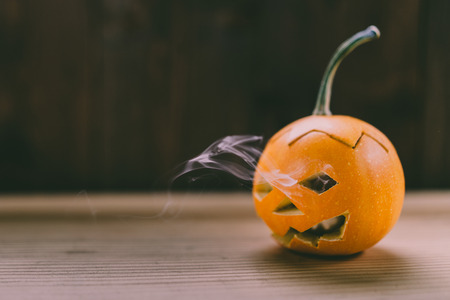 Closeup Photo of Carved Baby Pumpkin on the Wodden Background with Smokes Coming out of its Eyes and...