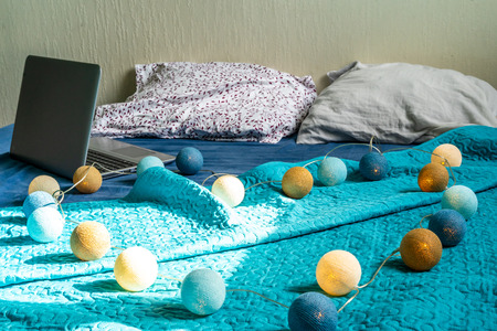 Grey Laptot Laying on Mattress with Turquoise Cover, Few Pillows and a White Background - with Christmas Decoration Lights on Bedの写真素材