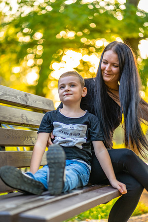 Kid Sitting on a Park Bench with His Mom  in Autum with Colorful Background in a Sunny Day - Caption on Shirt "I am, It`s now or never, Iask myself, Why I`m here"の写真素材