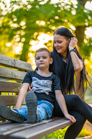 Kid Sitting on a Park Bench with His Mom  in Autum with Colorful Background in a Sunny Day - Caption on Shirt "I am, It`s now or never, Iask myself, Why I`m here"の写真素材