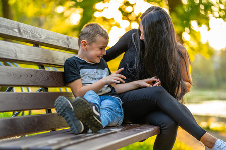 Mom Tickles Her Son on a Park Benck  in Autum with Colorful Backgroun in a Sunny Day, Both Laughing- Caption on Shirt "I am, It`s now or never, Iask myself, Why I`m here"の写真素材