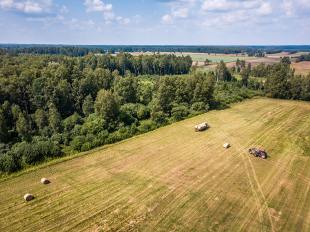 Aerial Drone Photo of Farmer Harvesting Hay Rolls in the Wheat Field with a Red Tractor - Sunny Summer Dayの写真素材