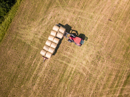 Aerial Drone Photo of Farmer Harvesting Hay Rolls in the Wheat Field with a Red Tractor - Sunny Summer Dayの写真素材