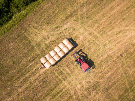 Aerial Drone Photo of Farmer Harvesting Hay Rolls in the Wheat Field with a Red Tractor - Sunny Summer Dayの写真素材