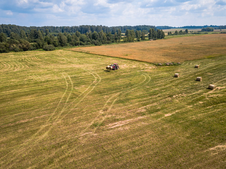 Aerial Drone Photo of Farmer Harvesting Hay Rolls in the Wheat Field with a Red Tractor - Sunny Summer Dayの写真素材