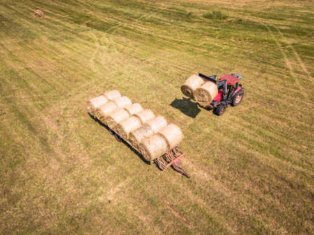 Aerial Drone Photo of Farmer Harvesting Hay Rolls in the Wheat Field with a Red Tractor - Sunny Summer Dayの写真素材