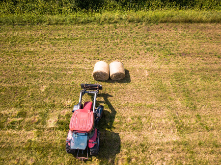 Aerial Drone Photo of Farmer Harvesting Hay Rolls in the Wheat Field with a Red Tractor - Sunny Summer Dayの写真素材