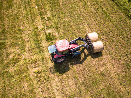 Aerial Drone Photo of Farmer Harvesting Hay Rolls in the Wheat Field with a Red Tractor - Sunny Summer Dayの写真素材