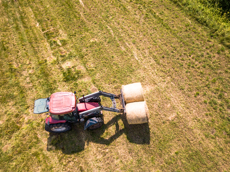 Aerial Drone Photo of Farmer Harvesting Hay Rolls in the Wheat Field with a Red Tractor - Sunny Summer Dayの写真素材