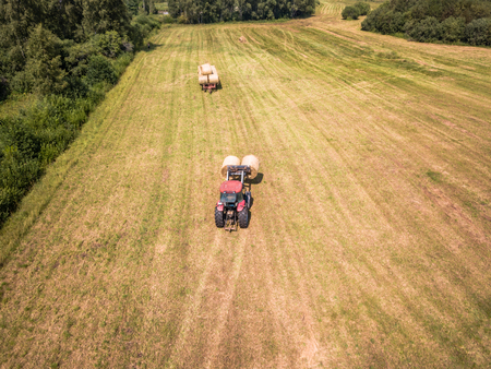 Aerial Drone Photo of Farmer Harvesting Hay Rolls in the Wheat Field with a Red Tractor - Sunny Summer Dayの写真素材