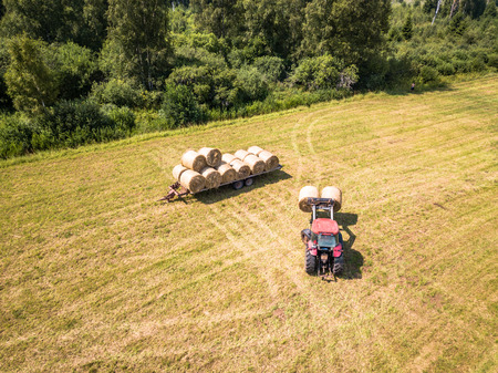 Aerial Drone Photo of Farmer Harvesting Hay Rolls in the Wheat Field with a Red Tractor - Sunny Summer Dayの写真素材