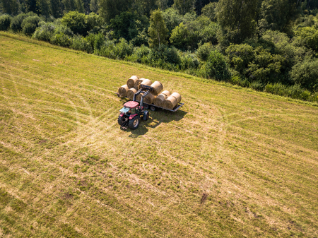 Aerial Drone Photo of Farmer Harvesting Hay Rolls in the Wheat Field with a Red Tractor - Sunny Summer Dayの写真素材