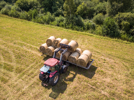 Aerial Drone Photo of Farmer Harvesting Hay Rolls in the Wheat Field with a Red Tractor - Sunny Summer Dayの写真素材
