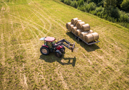 Aerial Drone Photo of Farmer Harvesting Hay Rolls in the Wheat Field with a Red Tractor - Sunny Summer Dayの写真素材
