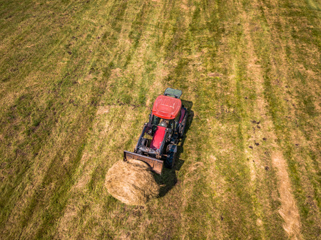 Aerial Drone Photo of Farmer Harvesting Hay Rolls in the Wheat Field with a Red Tractor - Sunny Summer Dayの写真素材