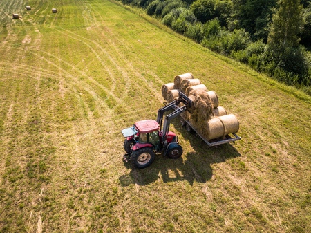 Aerial Drone Photo of Farmer Harvesting Hay Rolls in the Wheat Field with a Red Tractor - Sunny Summer Dayの写真素材