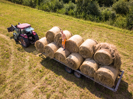 Aerial Drone Photo of Farmer Harvesting Hay Rolls in the Wheat Field with a Red Tractor - Sunny Summer Dayの写真素材