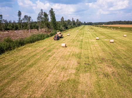 Aerial Drone Photo of Farmer Harvesting Hay Rolls in the Wheat Field with a Red Tractor - Sunny Summer Dayの写真素材