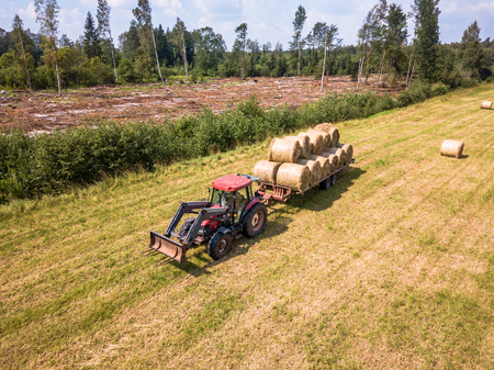 Aerial Drone Photo of Farmer Harvesting Hay Rolls in the Wheat Field with a Red Tractor - Sunny Summer Dayの写真素材
