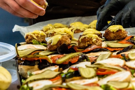 Female and Male Chef Putting Ingredients of Burgers on a Sliced Bread Spread on a Table in Black Gloves - Concept of the Hard Working Person and the Hygiene in the Kitchen, Partly Blurred Foregroundの写真素材