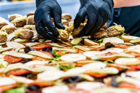 Female and Male Chef Putting Ingredients of Burgers on a Sliced Bread Spread on a Table in Black Gloves - Concept of the Hard Working Person and the Hygiene in the Kitchen, Partly Blurred Foregroundの写真素材