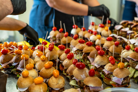 Female and Male Chef Putting Ingredients of Burgers on a Sliced Bread Spread on a Table in Black Gloves - Concept of the Hard Working Person and the Hygiene in the Kitchen, Partly Blurred Foregroundの写真素材