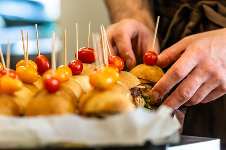 Male Chef Putting Ingredients of Burgers on a Sliced Bread Spread on a Table in Black Gloves - Concept of the Hard Working Person and the Hygiene in the Kitchen, Partly Blurred Foregroundの写真素材
