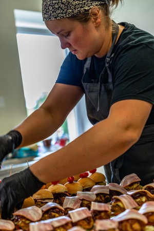 Female Chef Putting Ingredients of Burgers on a Sliced Bread Spread on a Table in Black Gloves - Concept of the Hard Working Person and the Hygiene in the Kitchen, Partly Blurred Foregroundの写真素材